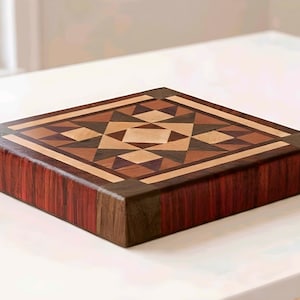 May include: Square wooden cutting board with a geometric design. The top features a mosaic pattern of various wood tones, including light beige, dark brown, and reddish-brown. The sides of the board are a mix of reddish-brown and dark brown wood.