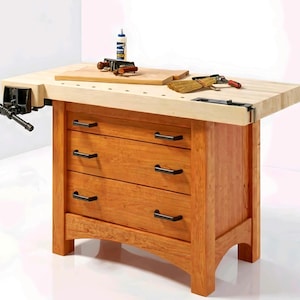 May include: A wooden workbench with a light-colored top and a three-drawer cabinet base. The drawers have black handles. A vise is attached to the left side of the workbench. Various tools and a cutting board are on the top.