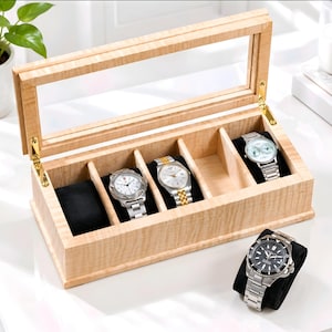 May include: A light-colored wooden watch display box with a hinged glass lid and gold-tone metal hinges. The interior features five individual compartments lined with black velvet cushions, three of which hold silver-tone wristwatches.