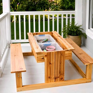 May include: A wooden sandbox table with two attached benches. The table has a rectangular design with two plastic bins inside, filled with sand and toys. The benches are made of the same light-colored wood. The table is on a white porch.