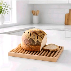May include: A round, golden-brown loaf of sourdough bread with a scored top sits on a rectangular wooden bread board with a slatted surface. One slice of the bread is cut and placed next to the main loaf on the light-colored wooden slats.