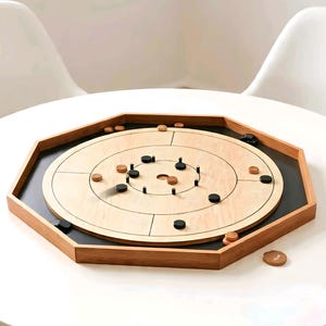 Puede incluir: Un juego de mesa carrom de madera con una superficie de juego de madera clara y un borde de madera oscura. El tablero tiene un área de juego circular con piezas de juego negras y marrones esparcidas por él. El tablero está sobre una mesa blanca.