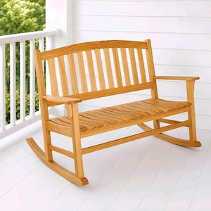 Puede incluir: Un banco mecedora de madera de color marrón claro con un respaldo curvo de listones verticales, reposabrazos planos y un asiento contorneado. El banco se apoya sobre dos balancines curvos conectados por travesaños horizontales en cada lado.