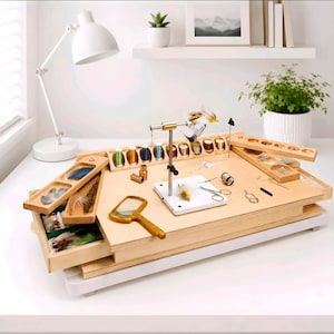 May include: A light-coloured wooden fly tying station featuring a central metal vice, multiple compartments for organising colourful threads, feathers, and various small metal tools, including a handheld magnifying glass and scissors.