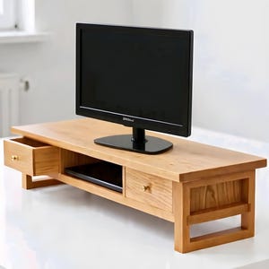 Peut inclure: Un meuble TV en bois brun clair avec deux tiroirs et une étagère. Un téléviseur à écran plat noir est placé dessus. Les tiroirs ont des boutons dorés. Le meuble est en bois massif.