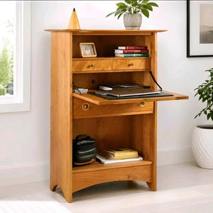 May include: A light brown wooden secretary desk featuring a drop-down work surface, two upper drawers, and one lower drawer. The desk includes open shelving, a flat work area with a black mat, and a storage compartment at the bottom.