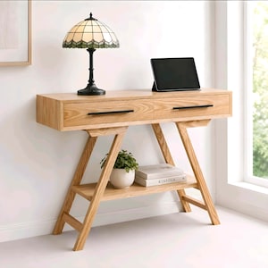 May include: A light-toned wooden console table featuring two drawers with thin black horizontal handles, an A-frame base, and a lower shelf. On the table sits a stained-glass lamp and a tablet. A small potted plant and a book are on the shelf.