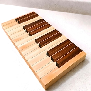May include: A wooden cutting board designed to resemble a piano keyboard. The board features alternating light and dark wood keys, with the dark keys being a rich brown color. The light keys are a natural wood tone.