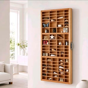 May include: A tall, rectangular wooden display case with numerous small, square compartments mounted on a wall. The shelves contain various decorative items including seashells, a blue butterfly, dominoes, wooden stars, and small metal objects.
