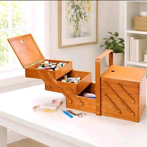 Puede incluir: Una caja de costura de madera con múltiples compartimentos abiertos, que revelan carretes de hilo y otros suministros de costura. La caja tiene un asa y está sobre una mesa blanca. También se ven una cinta métrica, tijeras y otras herramientas.