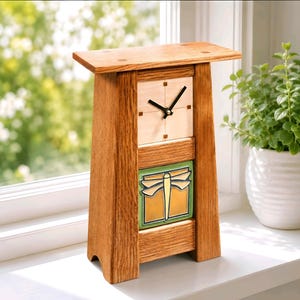 May include: A wooden mantel clock with a light-colored square face, black hands, and a decorative tile featuring a yellow dragonfly on a green background. The clock has a flared base and a flat top, constructed from medium-brown wood with visible grain.