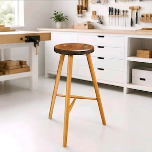 May include: A wooden stool with a round, dark brown seat and three light-coloured, tapered legs connected by a central support structure. The stool features a smooth, polished finish on the seat and a sturdy, functional design for seating.