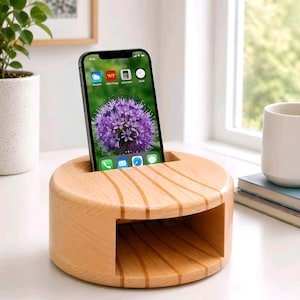 Op de afbeelding: Een lichtgekleurde houten telefoonstandaard met een sleuf om een smartphone in te plaatsen. De telefoon toont een paarse bloemafbeelding. De standaard heeft een cirkelvormig ontwerp met een rechthoekige opening.