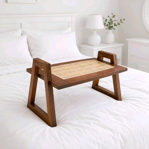 May include: A dark brown wooden bed tray with a woven rattan centre. The tray has a rectangular shape with handles and is placed on a white bed. The tray is designed for serving food or holding items. The bed is covered with white bedding and pillows.