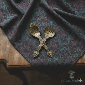 Puede incluir: Cuchara y tenedor de latón ornamentados cruzados sobre una tela estampada en verde azulado y burdeos. La cubertería detallada descansa sobre una mesa de madera, con la tela drapeada sobre ella. La tela tiene un diseño floral.