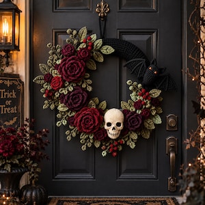 Puede incluir: Una corona circular con elementos de ganchillo que incluyen rosas de color rojo oscuro y burdeos, hojas verdes, pequeñas bayas rojas, un murciélago negro con ojos naranjas y una calavera central de color crema en una puerta negra.