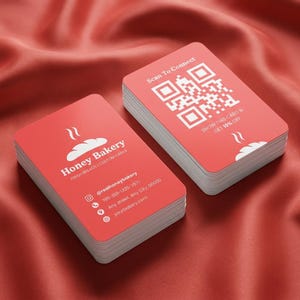 May include: Piles of red business cards with rounded corners. The cards display the text "Honey Bakery" with a bread graphic, contact details, and a QR code. The background is a red fabric.