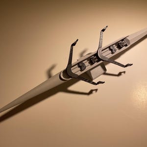 May include: A light gray model of a racing shell boat, viewed from above. The boat has a long, slender hull with a pointed bow and stern. The rigging is dark gray and includes oars and a sliding seat. The boat is on a light brown surface.