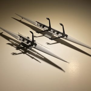 May include: Two gray model boats with black details are positioned on a light surface. The boats have long, pointed hulls and feature intricate black structures. The shadows cast by the boats add depth to the image.