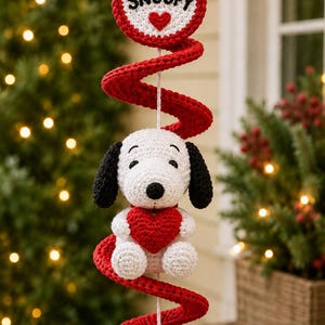 May include: A crocheted hanging decoration featuring a white Snoopy figure with black ears holding a red heart. Above the figure is a circular red and white sign with the text SNOOPY and a red heart. A red spiral crochet element surrounds the figure.