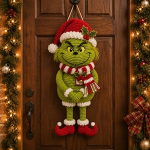 May include: A crocheted green character wall hanging wearing a red and white Santa hat with holly, a red and white striped scarf with a small bell, and red curled-toe shoes. The figure has a wide grin, yellow eyes, and is suspended by a twine loop.