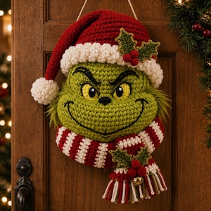 Op de afbeelding: Een gehaakte wandhanger met een groen karaktergezicht met gele ogen, een brede grijns en een rode kerstmuts met een witte pompon. Het personage draagt een rood-wit gestreepte sjaal versierd met groene hulstblaadjes en rode bessen.
