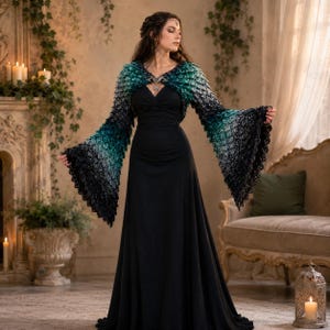 Puede incluir: Un vestido negro largo hasta el suelo con escote corazón y un bolero de mangas largas acampanadas. El bolero presenta un patrón degradado que va del verde azulado al negro con un diseño de ganchillo texturizado tipo escama.