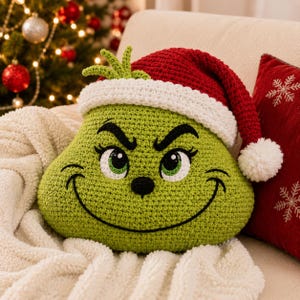 May include: A lime green crocheted pillow shaped like a character face with a wide black embroidered smile, green eyes, and a small black nose. The pillow wears a red and white crocheted Santa hat with a white pom-pom on the end of the hat.