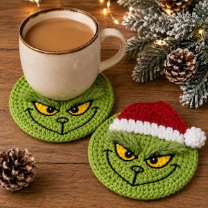 Puede incluir: Dos posavasos redondos de ganchillo verde con ojos amarillos bordados, cejas negras cosidas y una amplia sonrisa. Un posavasos incluye un gorro de Papá Noel de ganchillo rojo y blanco con un pompón blanco. Una taza de cerámica descansa sobre uno.