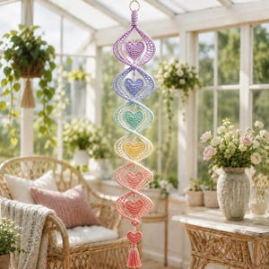 May include: A vertical hanging decoration featuring a spiral structure made of crocheted yarn in a rainbow gradient. The design includes seven small, solid crocheted hearts centred within each spiral loop, finished with a tassel at the bottom.