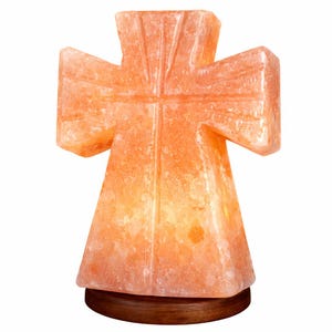 May include: A cross-shaped Himalayan salt lamp in shades of orange and pink. The lamp sits on a round, dark brown wooden base. The cross design features a central vertical line and horizontal arms, with a textured surface.