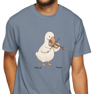 May include: A light blue t-shirt featuring a cartoon duck playing a violin. The duck is white with an orange beak and is holding a brown violin. The word "Violin" is printed below the duck.