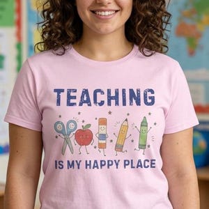 May include: A light pink short-sleeved t-shirt featuring the text "TEACHING IS MY HAPPY PLACE" in dark blue letters, accompanied by colourful illustrations of scissors, an apple, a glue stick, a pencil, and a crayon with small star accents.