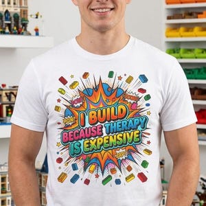 Könnte beinhalten: Ein weißes Kurzarm-T-Shirt mit einer bunten Grafik im Comic-Stil und dem Text "I BUILD BECAUSE THERAPY IS EXPENSIVE", umgeben von illustrierten Bausteinen und sternförmigen Elementen in Rot, Blau, Gelb und Grün.