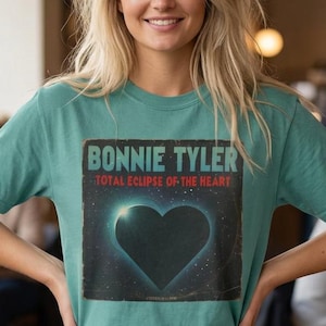 Puede incluir: Una camiseta de manga corta color verde azulado con un estampado gráfico de un corazón negro sobre un fondo de espacio estrellado. El texto BONNIE TYLER está impreso en azul claro sobre el corazón, con TOTAL ECLIPSE OF THE HEART en rojo debajo.