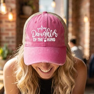 Pode incluir: Boné de caminhoneiro rosa e bege com o texto branco "Daughter of the King". O boné tem um visual desgastado e uma aba curva. O boné é usado por uma pessoa com cabelo loiro.