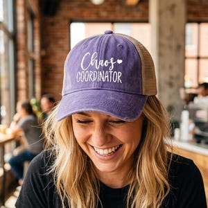 Pode incluir: Boné de caminhoneiro roxo e bege com as palavras "Chaos Coordinator" em escrita branca. O boné tem uma aba curva e uma parte traseira de malha. A pessoa que o usa está sorrindo.