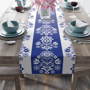 May include: A blue and white floral patterned table runner. The runner features a central blue stripe with white floral designs and white side stripes. The table is set with dishes, glasses, and flowers.