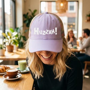 Pode incluir: Um boné de beisebol lilás com a palavra "Huzzah!" em letras brancas. O boné tem uma parte traseira de malha com um padrão listrado multicolorido. A pessoa que usa o boné está sorrindo, com uma xícara de café e um pastel na mesa.