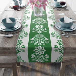 May include: A green and white floral patterned table runner. The runner is centered on a wooden table, with place settings and wine glasses on either side. A vase of pink flowers sits in the center.