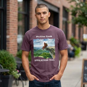 Puede incluir: Una camiseta granate con una ilustración escénica de McAfee Knob en el sendero de los Apalaches. El gráfico incluye el texto "McAfee Knob" y "Appalachian Trail". La camiseta es de manga corta y tiene cuello redondo.