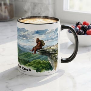 May include: White ceramic coffee mug with a black handle and rim. The mug features a watercolor illustration of a Bigfoot sitting on a cliff overlooking a mountain range. The mug contains a latte with a heart design.