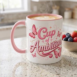 Pode incluir: Uma caneca de cerâmica branca com interior e alça cor-de-rosa, cheia de café com latte art. O exterior apresenta borboletas cor-de-rosa e o texto "Cup of Ambition" numa fonte cursiva rosa estilizada com espirais decorativas e pequenas estrelas.