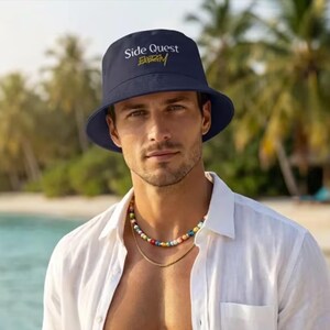 May include: Navy blue bucket hat with the text "Side Quest Energy" in white and gold. The hat is worn by a person wearing a white shirt and a colorful beaded necklace. The background is a beach scene.