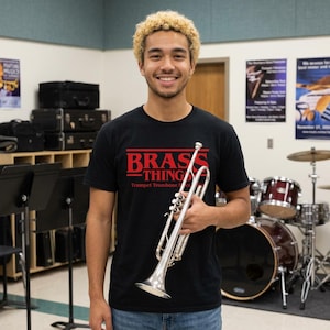 Peut inclure: Un t-shirt noir à manches courtes avec le texte rouge BRASS THINGS et les mots Trumpet Trombone Horn imprimés en dessous. Le t-shirt est en coton et est porté par une personne tenant une trompette argentée dans une salle de musique.
