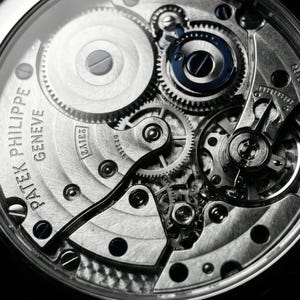 May include: Close-up of a watch mechanism with intricate gears and components. The watch face is visible, with the brand name "Patek Philippe Geneve" inscribed. The design features a mix of silver and blue elements.