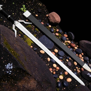 May include: A silver sword with a black handle and a matching black sheath. The sword is lying on a dark, textured surface with small rocks and pebbles. The handle has a decorative silver design.