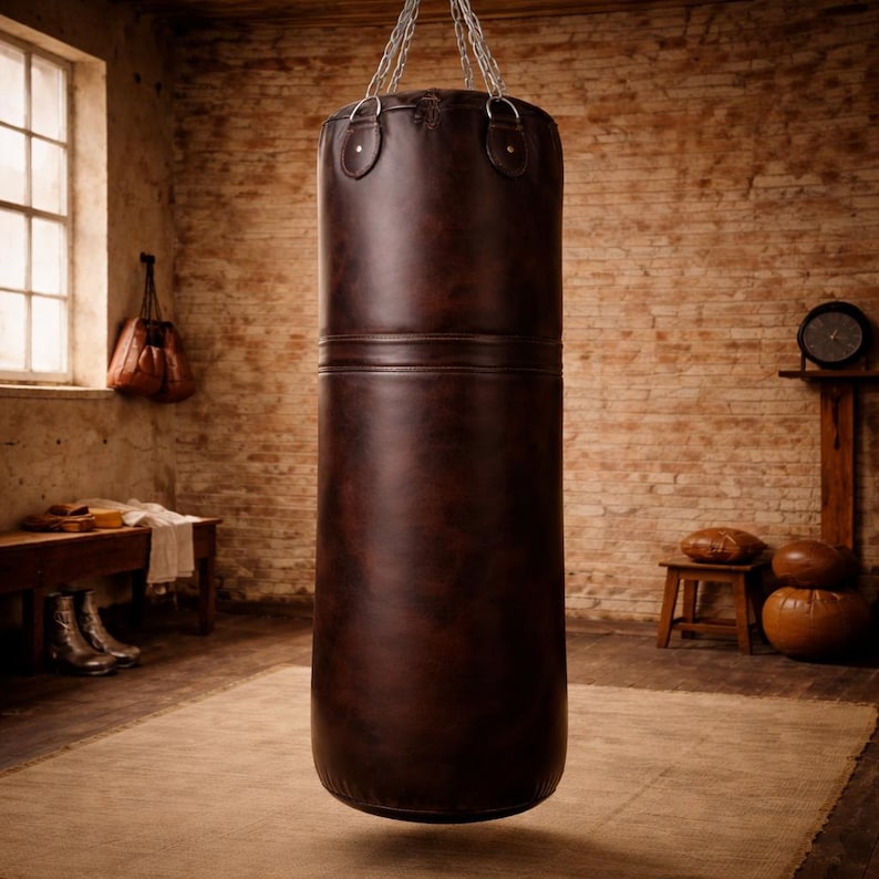 K&ouml;nnte beinhalten: Ein brauner Leder-Boxsack, der an einer Kette in einem Raum mit Backsteinmauern h&auml;ngt. Der zylindrische Sack hat eine horizontale Naht. Der Raum hat einen Teppich, eine Bank und ein Fenster.