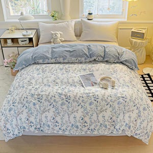 Puede incluir: Una cama con una funda nórdica con estampado floral azul y blanco y fundas de almohada a juego. La cama está hecha con un edredón azul claro y almohadas blancas. Un osito de peluche está encima de las almohadas. Una revista y unos auriculares están en la cama.
