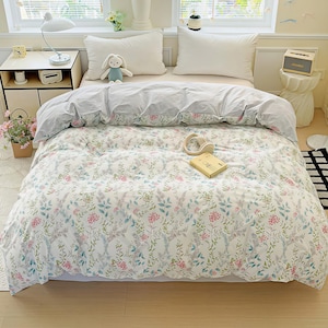 Puede incluir: Una escena de dormitorio con un juego de funda nórdica y almohadas con estampado floral. La ropa de cama presenta un fondo blanco adornado con un patrón de flores y follaje rosas, verdes y azules. Las almohadas blancas complementan el edredón, creando un ambiente relajante.